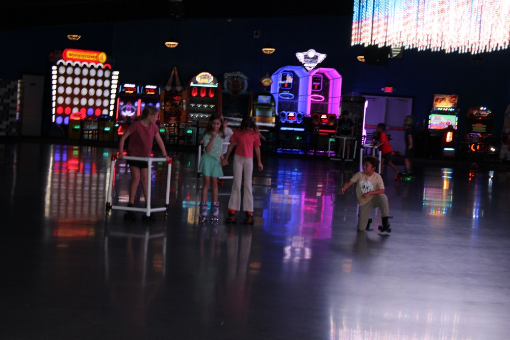 Children skate across a dimly lit roller rink in front of colorful arcade machines.